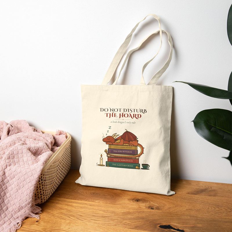 Natural canvas tote printed with a dragon asleep on books under Do Not Disturb the Hoard, on a wooden table