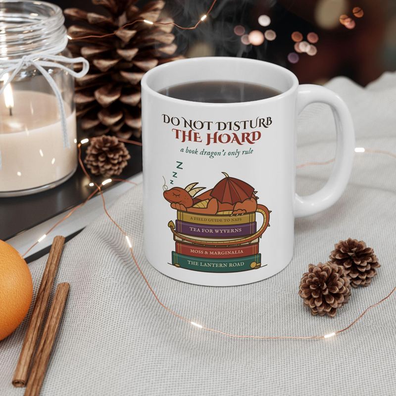 White mug printed with a copper dragon asleep on a stack of books under Do Not Disturb the Hoard, by pine cones