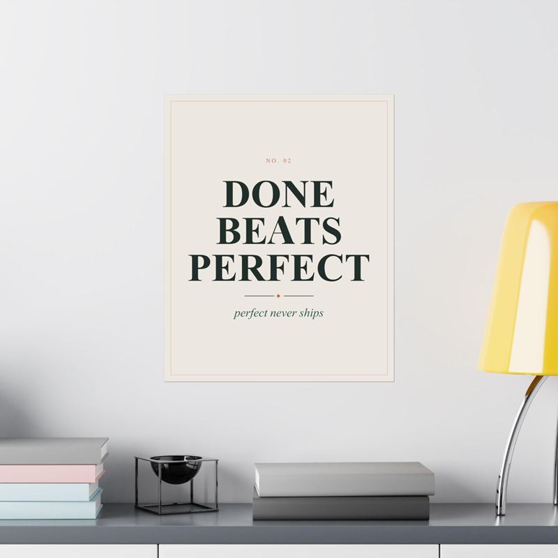 Cream poster reading Done Beats Perfect with "perfect never ships", on a white wall beside a yellow lamp