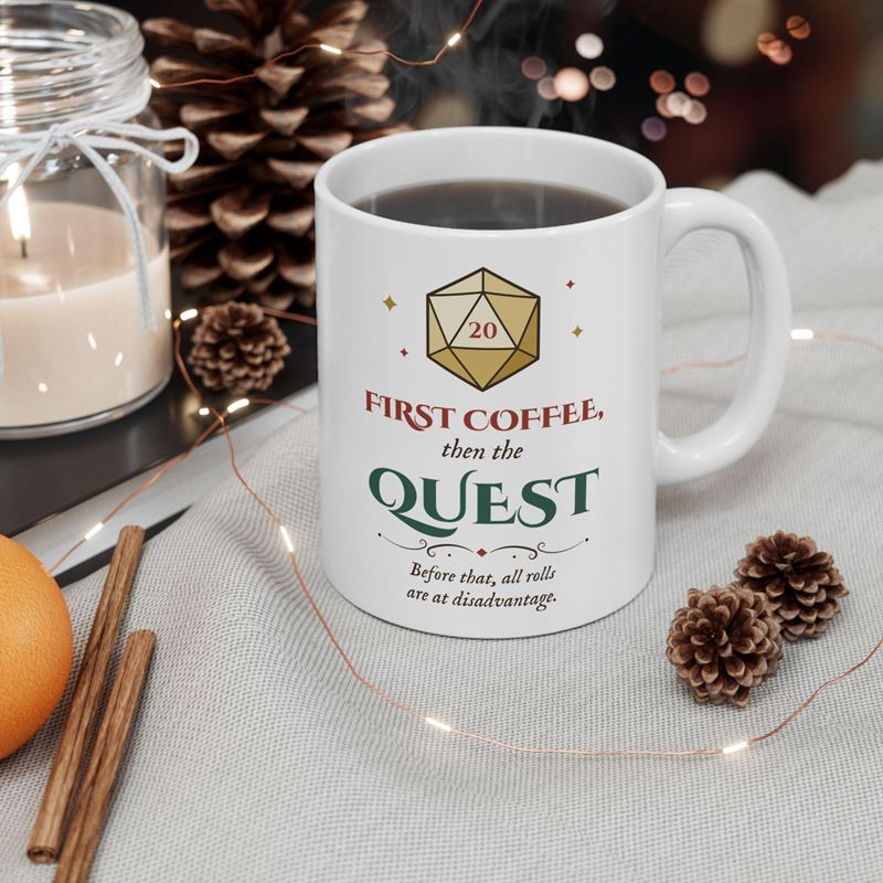 White mug printed with a gold D20 showing 20 and First Coffee, then the Quest, beside pine cones and cinnamon