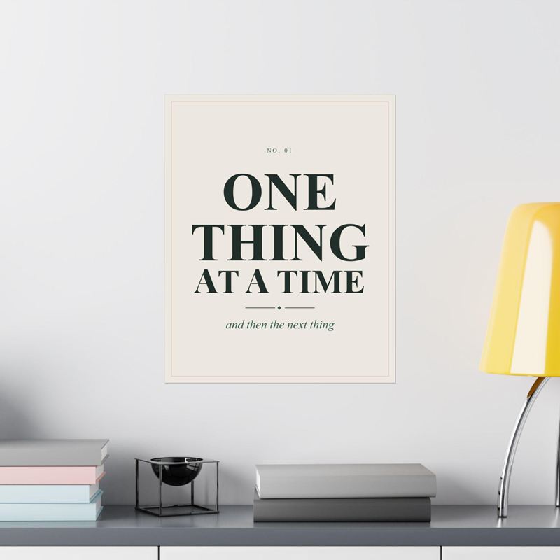 Cream poster reading One Thing at a Time with "and then the next thing", on a white wall beside a yellow lamp