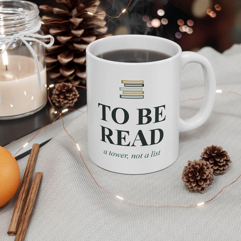 White mug printed with a small stack of books above To Be Read and "a tower, not a list", beside pine cones