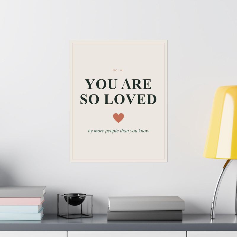 Cream nursery poster reading You Are So Loved with a terracotta heart, on a white wall beside a yellow lamp