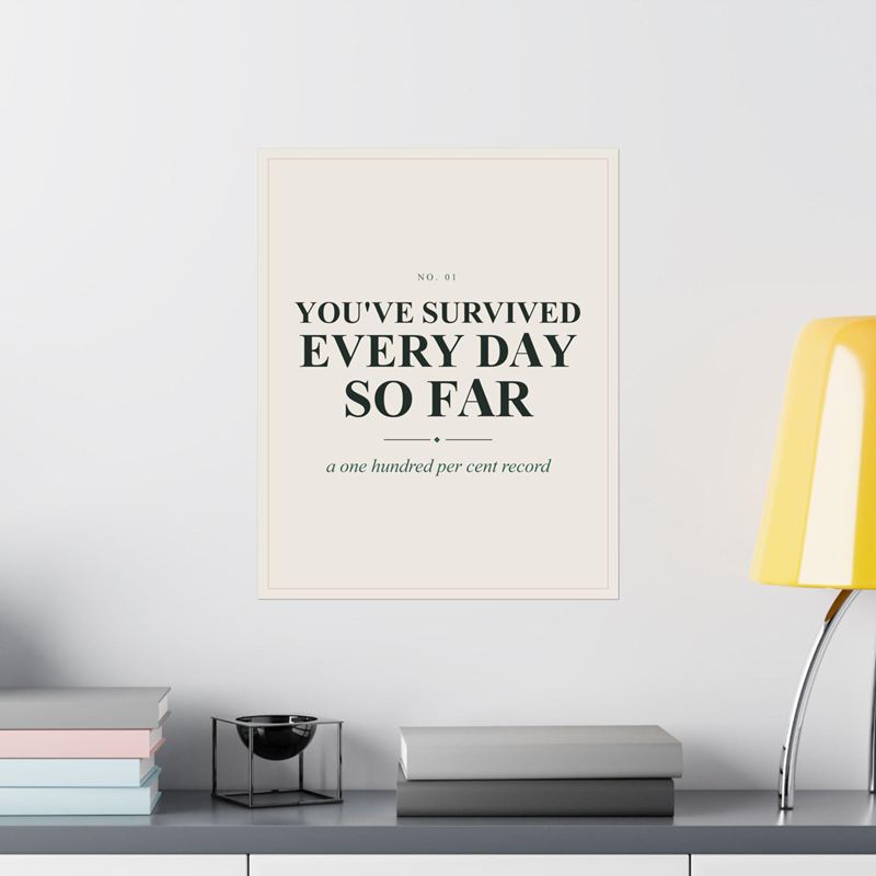 Cream poster reading You've Survived Every Day So Far, on a white wall above a shelf with a yellow lamp
